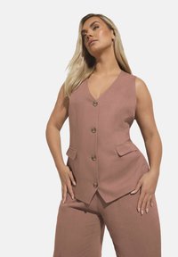 Gilet personalizzato di un rosa tenue con scollo a V, cinque bottoni e due tasche laterali, realizzato in tessuto testurizzato. Abbinato a pantaloni a gamba larga coordinati.