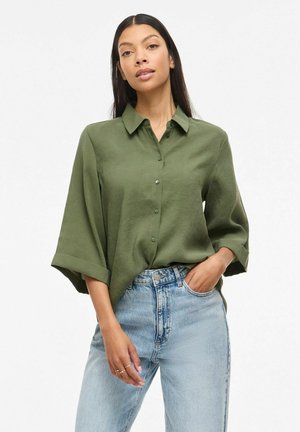 Femme portant un chemisier vert olive à boutons avec manches larges et un jean taille haute bleu clair, posant avec une main dans la poche.