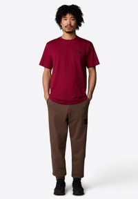 Czerwona koszulka z krótkim rękawem z czarnym logo na klatce piersiowej, zestawiona z brązowymi spodniami dresowymi z łatami na kolanach i naszywką z logo na udzie. Czarne sportowe buty.