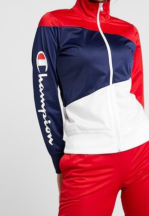 Femme portant une veste zippée rouge, blanche et bleue avec le logo "Champion" sur la manche et un pantalon rouge assorti.