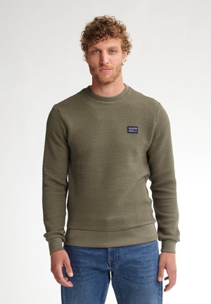 Felpa verde oliva con texture, colletto rotondo e polsini a coste, con una piccola toppa blu sul petto. Indossata con jeans blu.