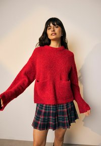 Rød chunky strikket sweater med lommer og brede ærmer, parret med en sort og rød ternet plisseret miniskirt. På toppen af designet og hyggelig tekstur.