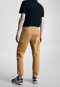 Tan cargobroek met elastische manchetten, twee voorzakken en twee zijzakken. Gecombineerd met een zwart shirt en sneakers.