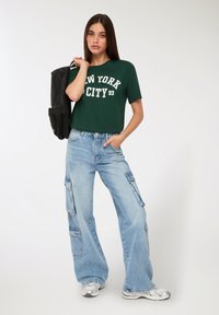 Giovane donna con t-shirt verde "New York City", jeans cargo azzurri chiari, sneakers argentati, che tiene uno zaino nero sulla spalla.