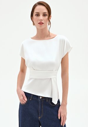 Donna che indossa una blusa bianca a maniche corte con una cintura larga in vita e jeans a vita alta blu scuro, in piedi con una mano in tasca.