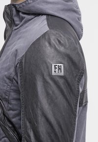 Grå jacka med en läderpanel på vänster axel och textilstruktur i grått. Märkbar hårdvara inkluderar en silverfärgad logotyp på ärmen.