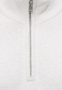 Gros plan d'un pull en maille blanche texturée avec une fermeture éclair argentée centrée, partiellement fermée.