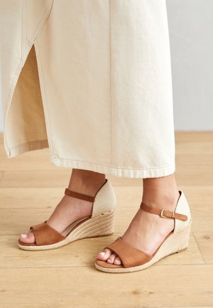Pieds portant des sandales compensées marron et beige avec brides à la cheville, associées à un pantalon large crème sur un sol en bois.