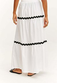 Weißer Maxirock mit gerafftem Design und drei schwarzen, wellenförmigen Ziernähten. Elastischer Bund. Kombiniert mit flachen schwarzen Sandalen.