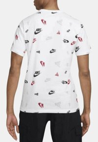 T-shirt bianco a maniche corte con un motivo ripetuto di vari logo Nike in nero, rosso e grigio. Collo a giro e vestibilità comoda.