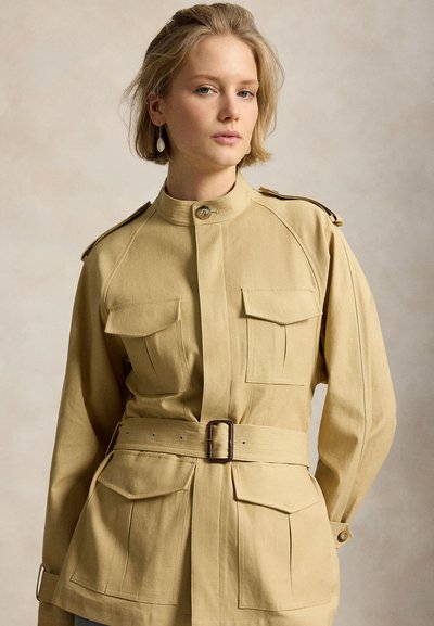 Femeie cu păr blond scurt purtând o jachetă de stil militar bej cu curea, cu buzunare la piept și talie, stând pe un fundal neutru.