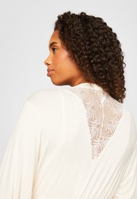 Cream-colored garment featuring a floral lace back detail, soft fabric, and flowing sleeves, with intricate lace patterns on the upper back.