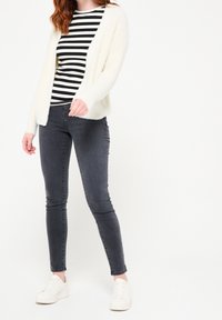 Vitt fluffigt cardigan över en svartvit randig topp, i kombination med mörkgrå skinny jeans och vita sneakers. Enkelt, avslappnat outfit.