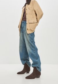Giacca beige con colletto marrone, sopra una maglietta marrone. Jeans azzurri che vanno dal chiaro allo scuro e stivali da cowboy in suede marrone scuro con tacco basso.