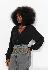 Blouse noire à manches bouffantes et col boutonné. Jean gris taille haute avec des accents brodés jaunes. Texture de tissu lisse.