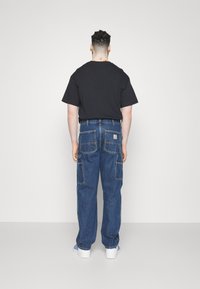 Denim-Cargo-Hosen in einer mittelblauen Waschung. Mit elastischen Bündchen, mehreren Gesäßtaschen und einem Logo-Patch hinten. Kombiniert mit einem schwarzen T-Shirt.