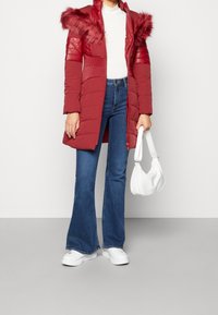Abrigo acolchado rojo con capucha de pelo sintético, detalles acolchados y cierre de cremallera. Combinado con jeans de campana azules y zapatillas blancas, sosteniendo un bolso blanco.