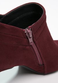 Burgundowe zamszowe botki za kostkę z bocznym zamkiem, o gładkiej teksturze i eleganckim kształcie. Wysoki obcas z okrągłym noskiem.