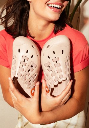 Mujer con camiseta coral sonriendo, sosteniendo un par de zapatos de lona tejida color beige claro con hebillas naranjas.