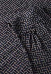 Bomullsskjorta har ett geometriskt mönster i marinblått på en ljusbrun bakgrund och har en rynkad axel samt en rundad nederkant.