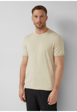 Mann mit kurzem Haar und Bart, der ein beigefarbenes kurzärmeliges Strickshirt und schwarze Hosen trägt, vor einem schlichten hellgrauen Hintergrund stehend.