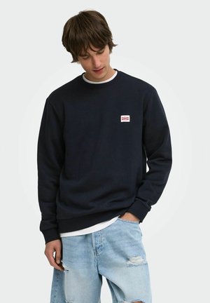 Jeune homme portant un sweat-shirt noir avec un petit logo rouge et blanc, jean clair déchiré, debout avec une main dans la poche, tête inclinée vers le bas.