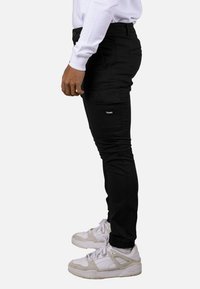 Pantalones cargo negros con diseño ajustado, múltiples bolsillos y textura suave, combinados con zapatillas blancas y una camisa blanca de manga larga.