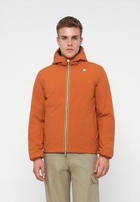 Mladý muž v oranžovej mikine s kapucňou a multicolor zipsom, oblečený v béžových cargo nohaviciach, postavený proti jednoduchému bielemu pozadiu.
