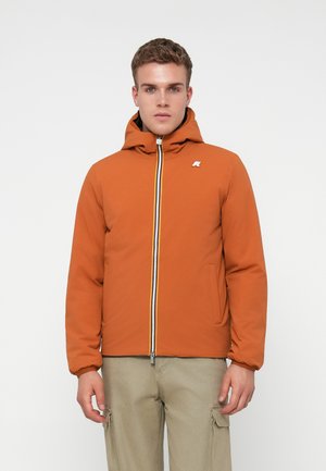 Un jeune homme portant une veste à capuche orange avec une fermeture éclair multicolore et un pantalon cargo beige, se tenant devant un fond blanc uni.