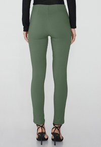 Zelené legíny na tělo vyrobené z elastického materiálu, s vysokým pasem a hladkou texturou, spárované s černými sandály na vysokém podpatku.