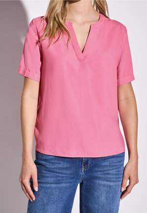 Vrouw draagt een roze blouse met korte mouwen en V-hals, gecombineerd met een blauwe spijkerbroek, staand tegen een lichte achtergrond.