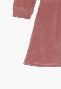 Dlouhý rukáv šaty v měkké, jemně růžové plyšové tkanině. Obsahují zakřivený lem a žebrované manžety, s hladkou texturou a minimálním šitím.