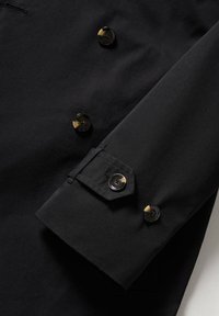 Manche et devant d'un manteau en tissu noir avec des boutons bicolores brillants, comportant un détail de sangle boutonnée sur le poignet.