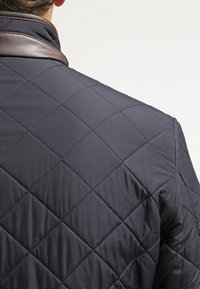 Gep quiltete marineblaue Jacke mit einem Rautenmuster, einem kontrastierenden braunen Lederkragen und einer sanften Textur auf dem Stoff.