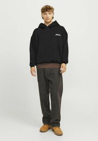 Zwarte oversized hoodie met een voorzak en witte logo. Gecombineerd met donkere spijkerbroeken en tan kleurige werklaarzen. Eenvoudige achtergrond.