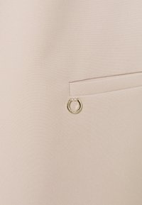 Tissu beige avec une finition texturée, présentant une poche discrète et un accent circulaire doré attaché près de l'ouverture.