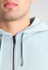 Sweat à capuche bleu clair avec fermeture éclair, cordon de serrage, tissu lisse, texture côtelée près de la fermeture éclair et poche kangourou en bas.