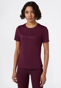Joven con cabello rizado corto que lleva una camiseta ajustada de color burdeos de Champion y leggings a juego, posando frente a un fondo blanco liso.