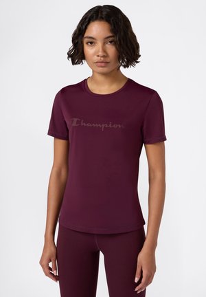Giovane donna con capelli corti e ricci, che indossa una t-shirt aderente color prugna della Champion e leggings abbinati, che si trova di fronte a uno sfondo bianco semplice.