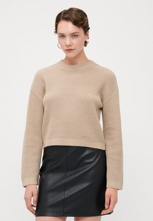 Beige knitted sweater with a crew neck and ribbed cuffs, paired with a black faux leather mini skirt. Smooth texture and casual fit.
