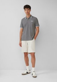 Grijze polo met een witte gestreepte kraag en manchetten, gecombineerd met lichtbeige shorts. Model draagt witte sneakers met zwarte accenten.