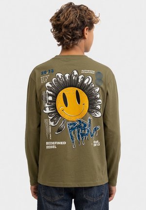 Personne portant une chemise à manches longues vert olive avec un grand motif d'une fleur jaune souriante et un texte dans le dos.