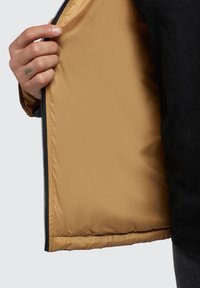Black zip-up jacket with a tan, smooth inner lining; hand holding the jacket's opening shows the zipper and stitching details.