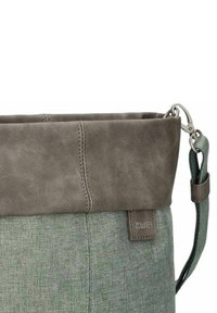 Bolsa de tela verde con detalles de cuero gris, que presenta un diseño de parte superior enrollable, correa ajustable y una etiqueta con el logotipo en el lateral. Superficies suaves y con textura.