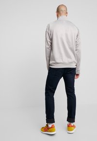 Lys grå zip-up jakke med en glat tekstur, ribbede manchetter og krave. Sammen med mørke jeans og farverige gule og røde sneakers.
