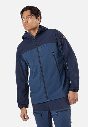 Giovane uomo che indossa una giacca con cappuccio blu navy con cerniera e pantaloni abbinati, che guarda verso sinistra su uno sfondo neutro.