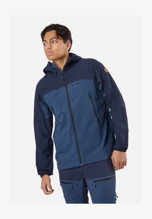 Junger Mann trägt eine marineblaue und blaue Kapuzenjacke mit Reißverschluss und passende Hose, schaut nach links vor einem einfarbigen Hintergrund.