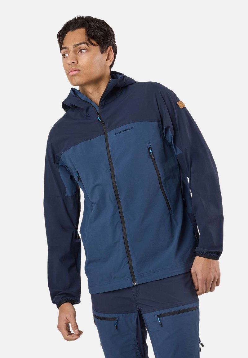 Nuori mies, joka pukeutuneena tummansiniseen ja siniseen vetoketjulliseen huppariin ja vastaaviin housuihin, katsoo vasemmalleen yksinkertaisen taustan edessä.