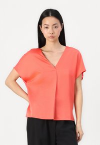 Koraļļu zīda blūze ar V-veida kakla izgriezumu, īsām piedurknēm un brīvu piegriezumu; ar centrālo krokām un gludu, spīdīgu tekstūru. Apvienota ar melnām biksēm.