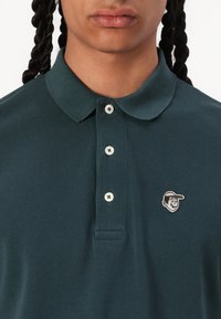 Mørkegrønn polo skjorte laget av teksturert stoff, med en klassisk krage, tre knapper og et sydd på tegneserieansiktslogo.